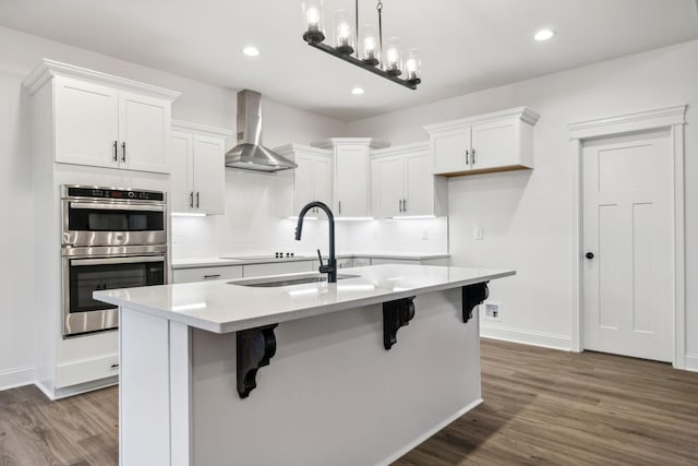 kitchen with white cabinetry, stainless steel double oven, wall chimney range hood, and an island with sink