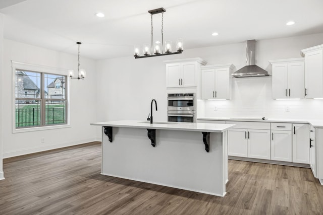 kitchen featuring wall chimney exhaust hood, light hardwood / wood-style flooring, double oven, a kitchen island with sink, and white cabinets