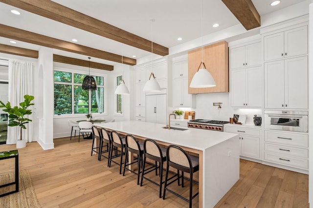 kitchen with beam ceiling, white cabinetry, hanging light fixtures, a kitchen island with sink, and light wood-type flooring