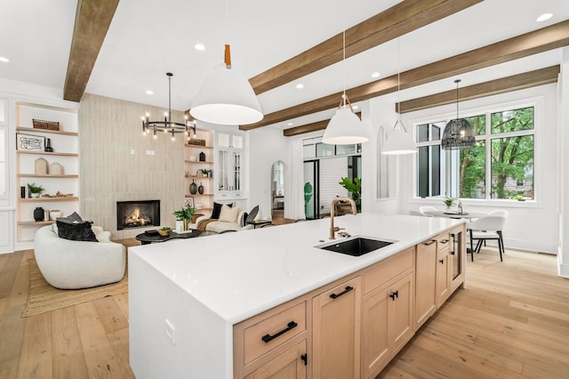 kitchen with light brown cabinetry, sink, beam ceiling, hanging light fixtures, and an island with sink