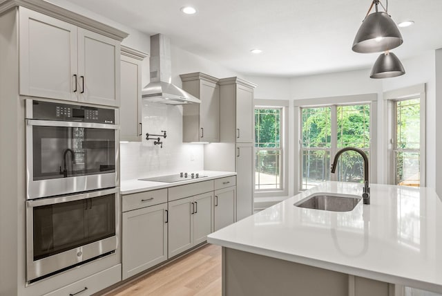 kitchen with sink, wall chimney range hood, double oven, light hardwood / wood-style floors, and black electric cooktop