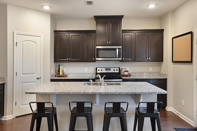 kitchen featuring dark brown cabinetry, dark wood-type flooring, light stone counters, a center island with sink, and appliances with stainless steel finishes