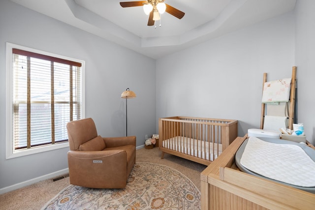 carpeted bedroom with a raised ceiling, ceiling fan, and a nursery area