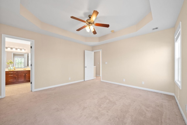 unfurnished bedroom with ensuite bathroom, a raised ceiling, ceiling fan, and light colored carpet