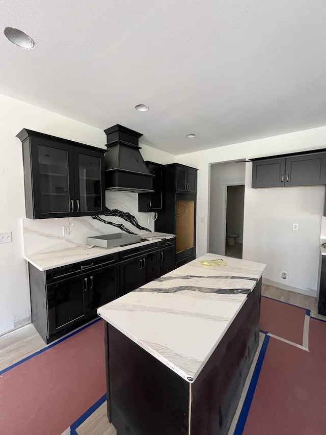 kitchen with glass insert cabinets, light stone counters, a center island, dark cabinetry, and premium range hood