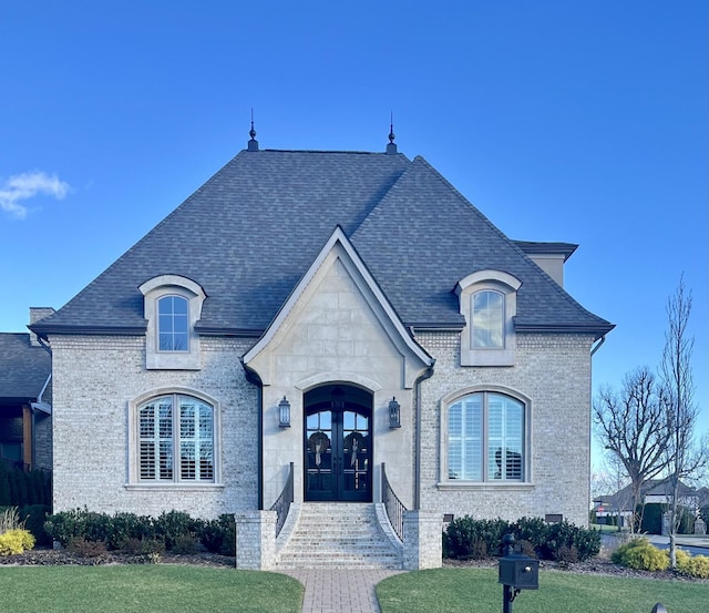 french country home featuring french doors, a front lawn, a shingled roof, and brick siding