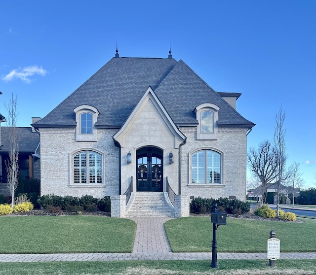 french country inspired facade featuring french doors, roof with shingles, a front yard, and brick siding