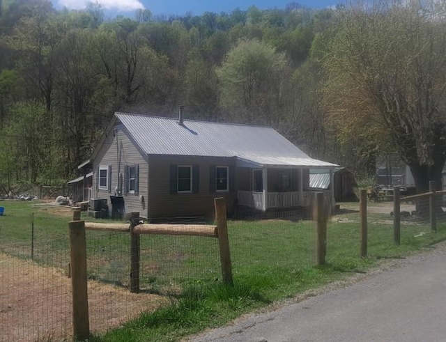 single story home with a front yard, central AC unit, metal roof, fence, and a forest view