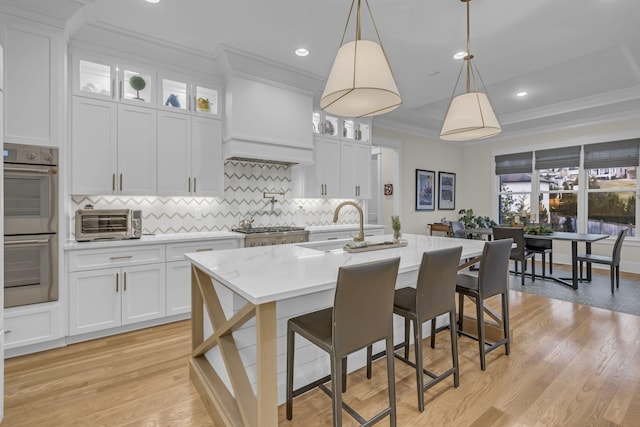 kitchen with custom range hood, a center island with sink, light hardwood / wood-style flooring, white cabinets, and hanging light fixtures