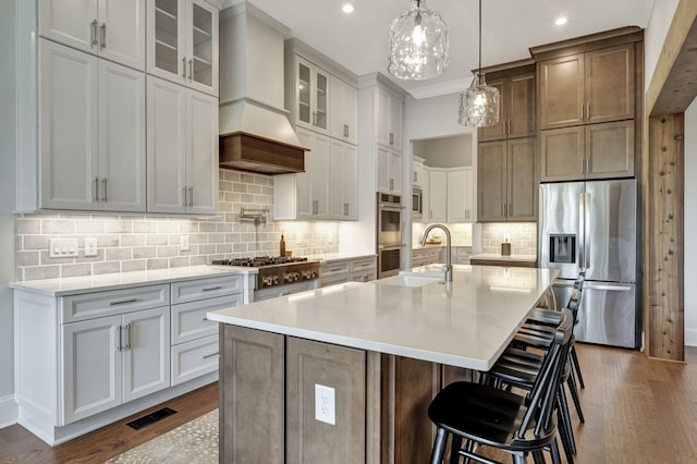 kitchen featuring sink, an island with sink, decorative light fixtures, custom range hood, and appliances with stainless steel finishes