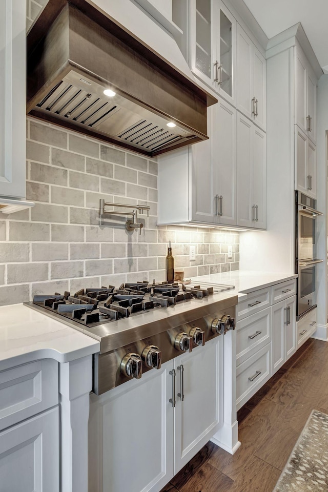 kitchen with white cabinetry, dark wood-type flooring, tasteful backsplash, custom range hood, and appliances with stainless steel finishes