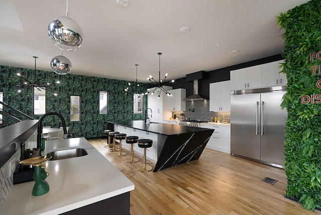 kitchen with white cabinetry, sink, stainless steel appliances, wall chimney range hood, and a center island with sink