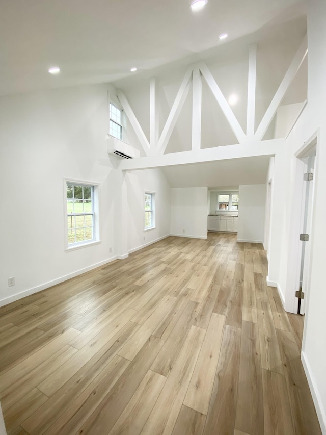 unfurnished living room with a healthy amount of sunlight, a wall mounted AC, high vaulted ceiling, and light wood-type flooring