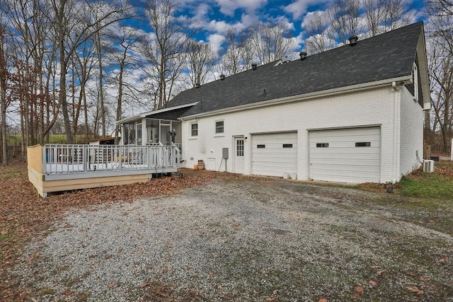 back of property featuring a sunroom, roof with shingles, gravel driveway, a wooden deck, and brick siding