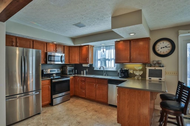 kitchen featuring a peninsula, a sink, appliances with stainless steel finishes, brown cabinetry, and dark countertops