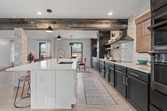 kitchen with sink, wall chimney range hood, light hardwood / wood-style flooring, black electric cooktop, and a center island with sink