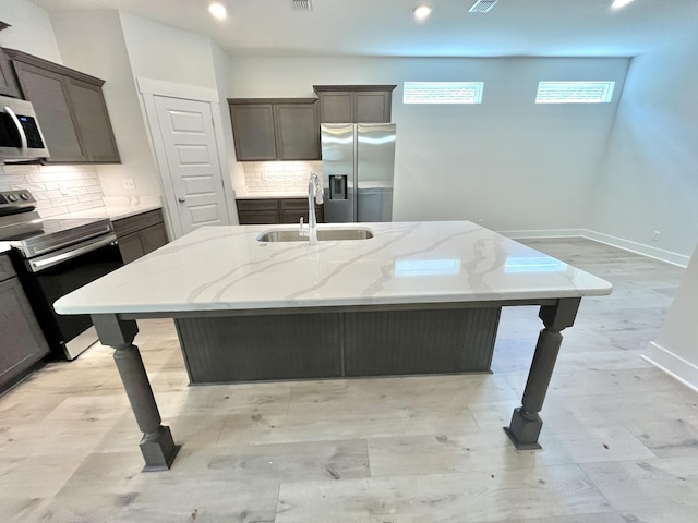 kitchen with a center island with sink, a sink, stainless steel appliances, light stone countertops, and dark brown cabinets