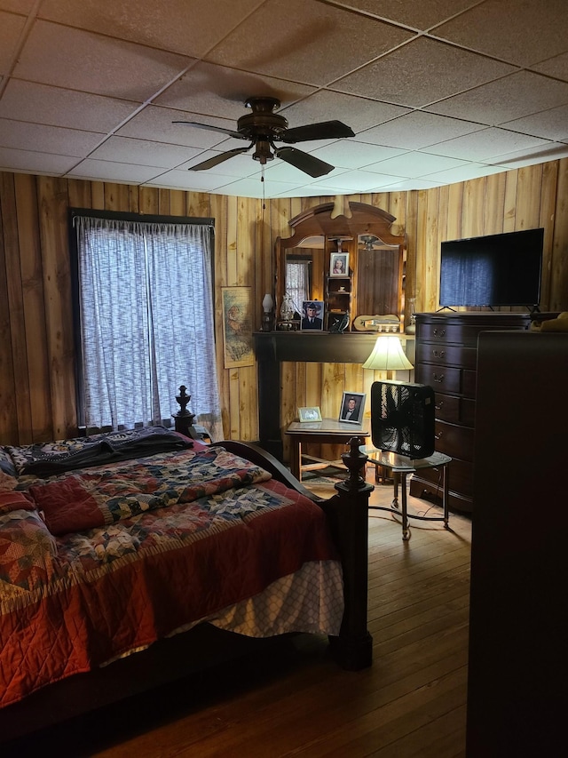 bedroom featuring hardwood / wood-style flooring, a drop ceiling, wood walls, and ceiling fan