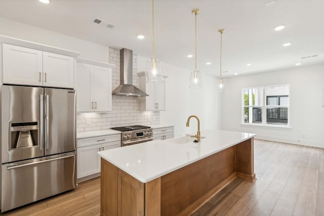 kitchen featuring a center island with sink, wall chimney range hood, sink, appliances with stainless steel finishes, and white cabinetry