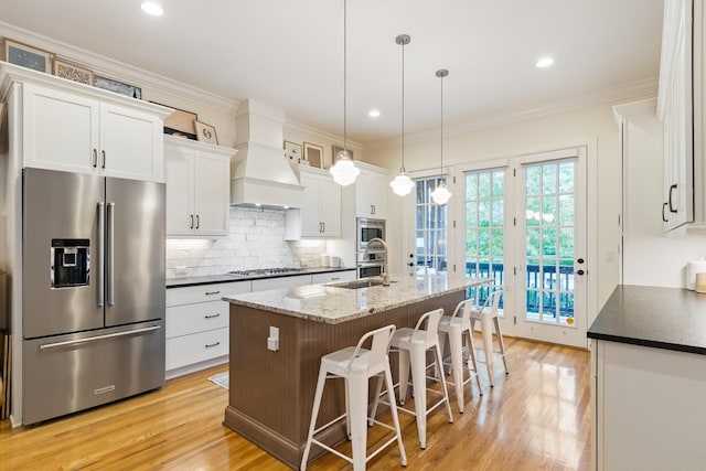 kitchen with white cabinetry, light hardwood / wood-style flooring, premium range hood, a center island with sink, and appliances with stainless steel finishes