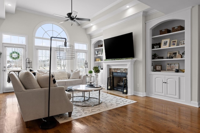 living room with built in features, a stone fireplace, hardwood / wood-style floors, and crown molding