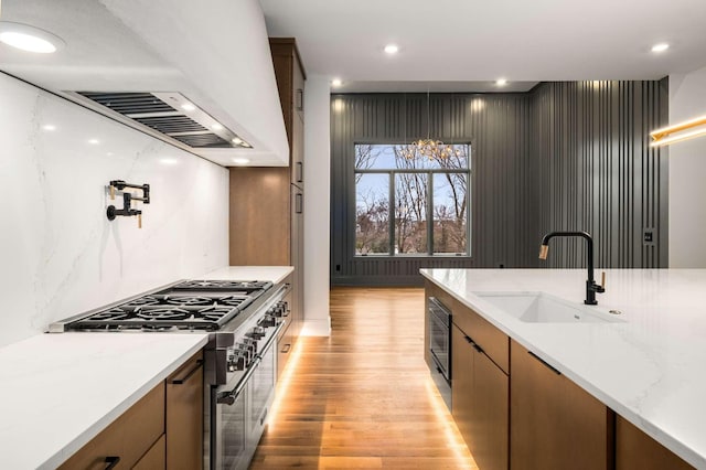 kitchen with exhaust hood, sink, high end stainless steel range, light stone countertops, and light hardwood / wood-style floors