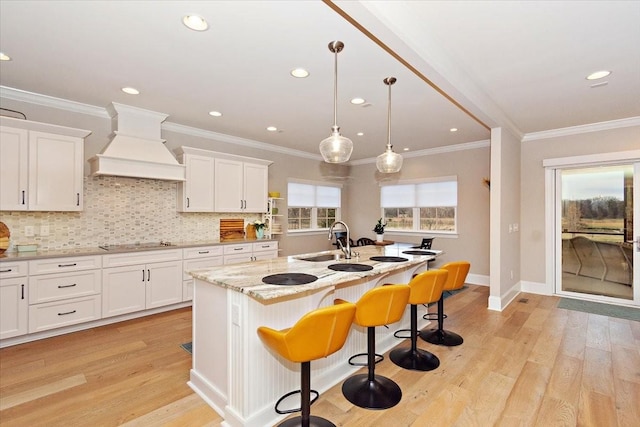 kitchen featuring pendant lighting, custom exhaust hood, a kitchen island with sink, white cabinets, and light hardwood / wood-style floors