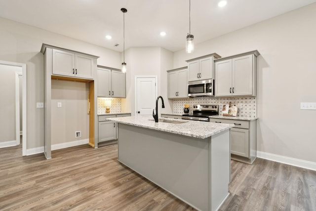 kitchen with light stone countertops, appliances with stainless steel finishes, pendant lighting, light hardwood / wood-style flooring, and an island with sink