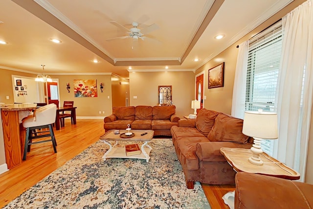 living room featuring ceiling fan with notable chandelier, light wood-type flooring, a raised ceiling, and ornamental molding