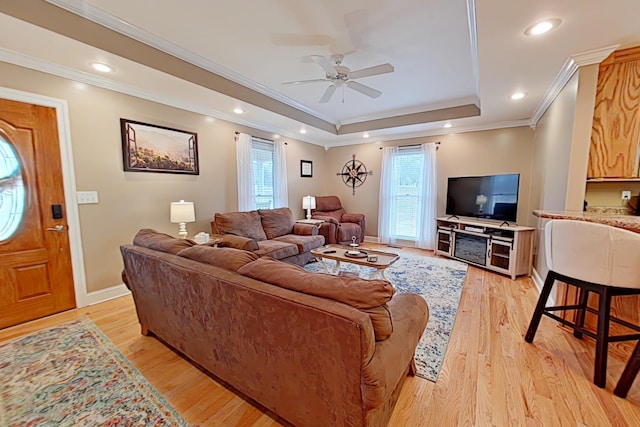 living room featuring a raised ceiling, ceiling fan, ornamental molding, and light wood-type flooring