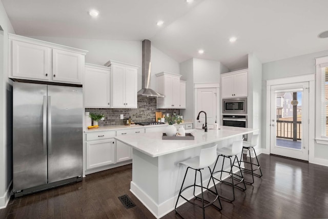 kitchen featuring a kitchen island with sink, white cabinets, wall chimney range hood, vaulted ceiling, and stainless steel appliances