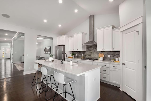 kitchen featuring appliances with stainless steel finishes, vaulted ceiling, wall chimney range hood, white cabinets, and an island with sink