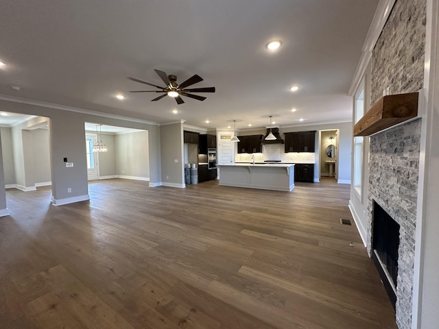 unfurnished living room with ceiling fan with notable chandelier, dark wood-type flooring, ornamental molding, and a stone fireplace