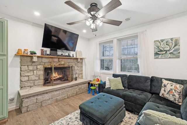 living room with ceiling fan, a stone fireplace, wood-type flooring, and crown molding