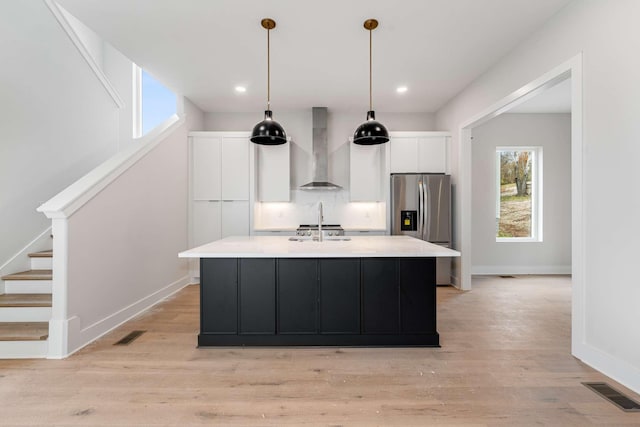 kitchen featuring white cabinetry, wall chimney range hood, stainless steel fridge, pendant lighting, and a kitchen island with sink