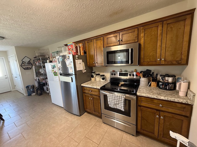 kitchen featuring a textured ceiling, stainless steel appliances, brown cabinetry, and light stone countertops
