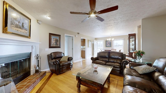 living room with ceiling fan with notable chandelier, a brick fireplace, light wood-type flooring, and a textured ceiling