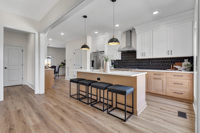 kitchen featuring a center island with sink, white cabinets, wall chimney range hood, hanging light fixtures, and appliances with stainless steel finishes