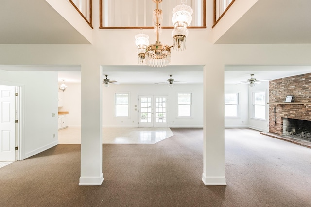 unfurnished living room featuring french doors, ceiling fan with notable chandelier, a brick fireplace, carpet flooring, and a towering ceiling