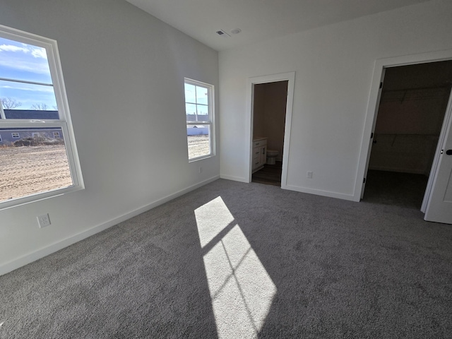 unfurnished bedroom featuring a spacious closet, ensuite bath, and dark colored carpet