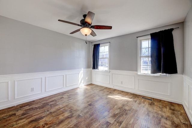 empty room with a ceiling fan, dark wood-style flooring, visible vents, and a wainscoted wall