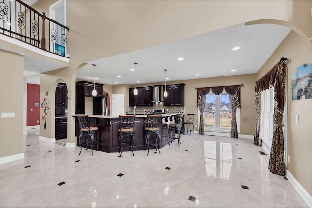 kitchen with decorative backsplash, wall chimney exhaust hood, dark brown cabinetry, a kitchen island with sink, and decorative light fixtures