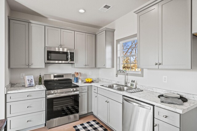 kitchen with light stone counters, gray cabinetry, stainless steel appliances, sink, and light hardwood / wood-style floors