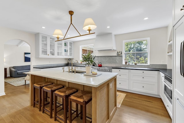 kitchen featuring custom exhaust hood, a kitchen island with sink, white cabinets, oven, and sink