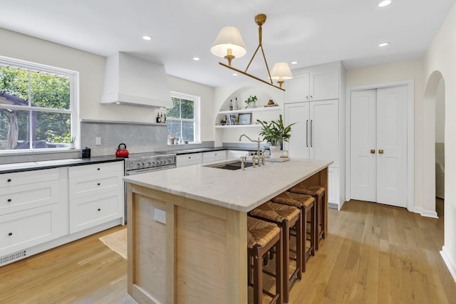 kitchen with premium range hood, sink, white cabinets, hanging light fixtures, and an island with sink