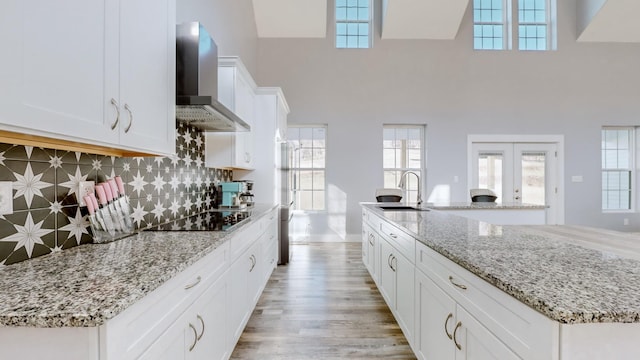 kitchen featuring a high ceiling, wall chimney range hood, decorative backsplash, black electric cooktop, and white cabinetry