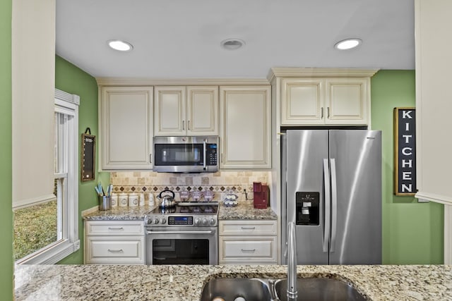 kitchen with decorative backsplash, light stone counters, cream cabinetry, and appliances with stainless steel finishes