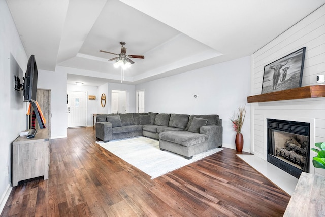 living room with ceiling fan, a large fireplace, a tray ceiling, and dark hardwood / wood-style flooring