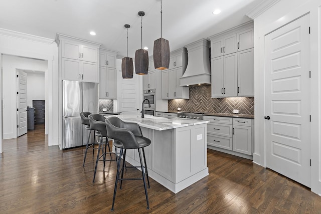 kitchen with a kitchen bar, premium range hood, a kitchen island with sink, pendant lighting, and stainless steel fridge