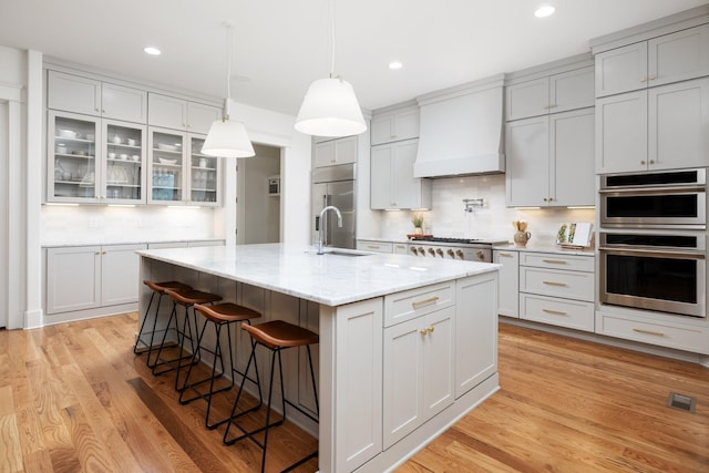 kitchen featuring backsplash, stainless steel appliances, custom range hood, an island with sink, and light wood-type flooring